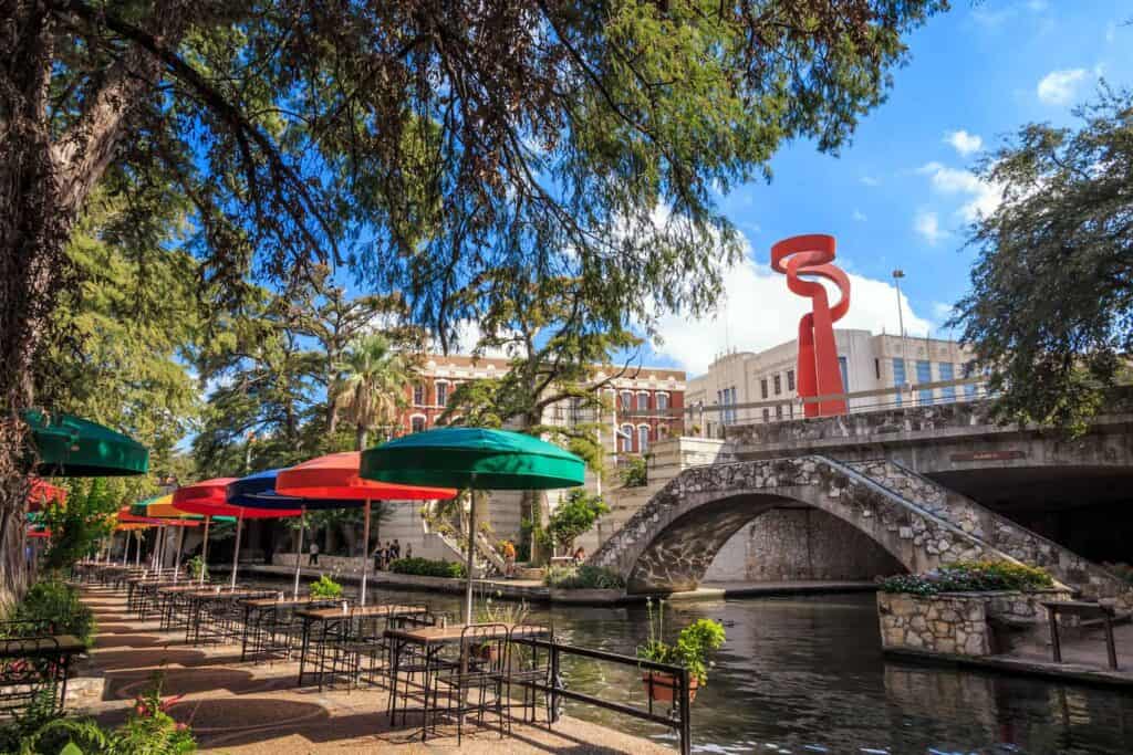 Une vue sur la promenade fluviale de San Antonio.  Il y a un ensemble de parasols colorés au-dessus des tables et un pont sur la photo. 